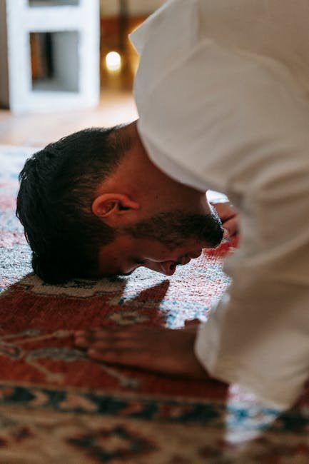 A person in traditional attire bows in prayer on a patterned rug within a room.
